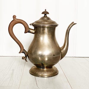 May include: A vintage silver-toned teapot with a brown wooden handle and a decorative lid. The teapot has a curved spout and sits on a detailed base. The background is a light-colored wooden surface.
