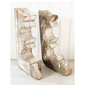 May include: Two distressed wooden corbels with a white painted finish. The corbels feature a decorative, curved design with multiple cut-out sections. The wood shows signs of age and wear, adding to their rustic charm and vintage appeal.