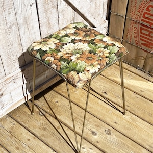 May include: Vintage stool with a square floral-patterned seat in shades of brown, green, and cream. The seat is supported by slender, gold-colored metal legs. The stool is placed on a wooden deck.
