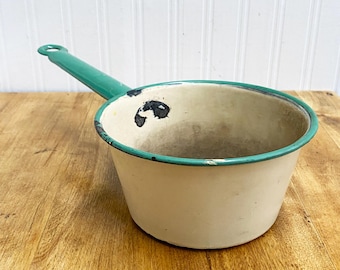 May include: A vintage white enamel saucepan with a teal green handle. The saucepan has a worn, distressed look with some black marks on the inside.