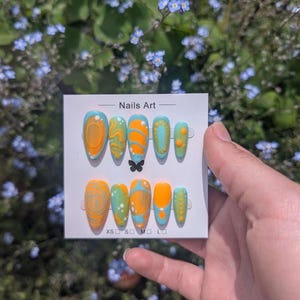 May include: A set of ten press-on nails displayed on a white card labelled Nails Art. The nails feature a vibrant colour palette of bright orange, light blue, and lime green with various abstract patterns, including swirls, dots, and raised textures.