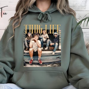 May include: Sage green hoodie with a graphic of four people on a porch, with the words "THUG LIFE" in gold lettering. The hoodie has a drawstring hood and a kangaroo pocket.