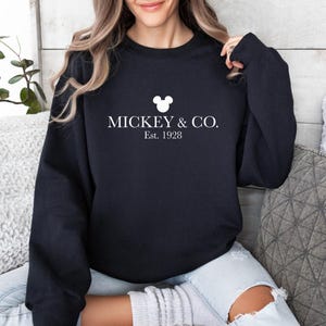 a woman sitting on a couch wearing a mickey and co sweatshirt