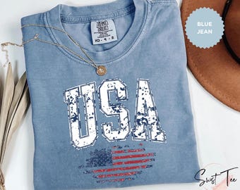 May include: A blue jean colored t-shirt with the word "USA" in distressed white lettering and a distressed American flag design. A gold necklace with a pendant is draped over the shirt. The shirt is a Comfort Colors brand.