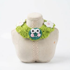 May include: A lime green crocheted collar featuring a central circular frog face with large white eyes and pink cheeks, accented by two white crocheted flowers on the sides. The collar has a scalloped edge and a textured, yarn-based construction.