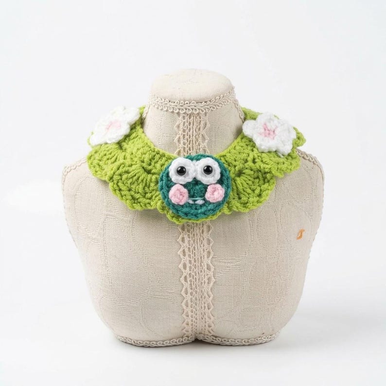 May include: A lime green crocheted collar featuring a central circular frog face with large white eyes and pink cheeks, accented by two white crocheted flowers on the sides. The collar has a scalloped edge and a textured, yarn-based construction.