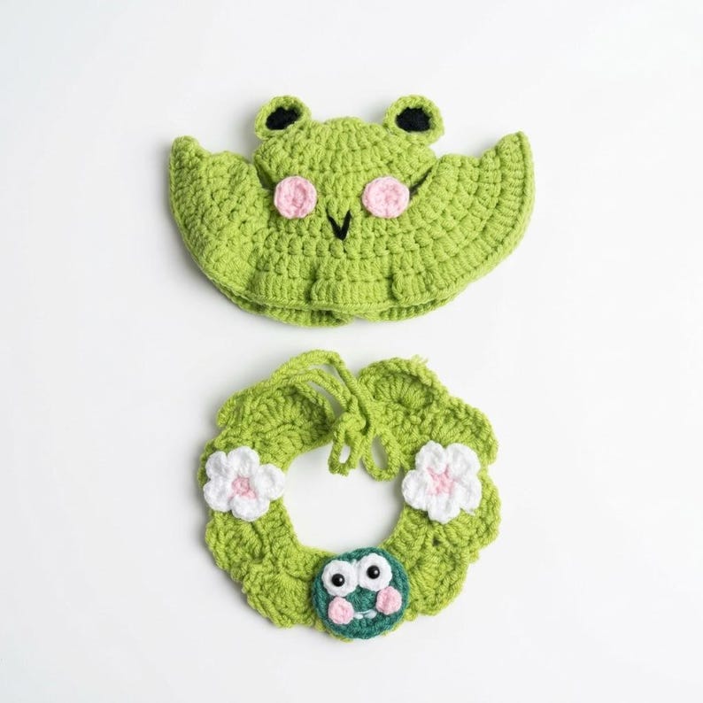 May include: A light green crocheted hat featuring a frog face with black eyes and pink cheeks, paired with a matching light green crocheted collar adorned with two white flowers and a small frog face with white eyes and pink cheeks.