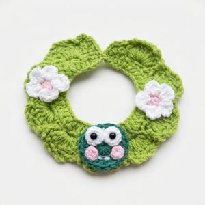 May include: A circular crochet accessory in bright green featuring a scalloped edge, two white flowers with light pink centres, and a central green frog face with large white eyes, black pupils, and light pink cheeks on a plain background.
