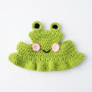May include: A bright green crocheted hat shaped like a frog, featuring two protruding eyes with black pupils, two small pink circular cheeks, and a simple black stitched mouth. The hat has a wide, ruffled brim and a textured yarn construction.