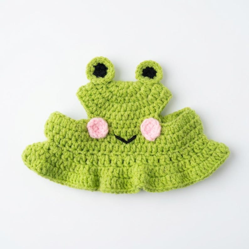 May include: A bright green crocheted hat shaped like a frog, featuring two protruding eyes with black pupils, two small pink circular cheeks, and a simple black stitched mouth. The hat has a wide, ruffled brim and a textured yarn construction.