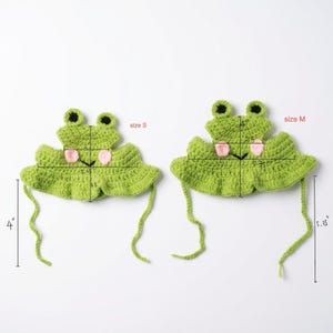 May include: Two green crochet frog hats with pink cheeks and black eyes. The size S hat measures 2 inches wide at the top, 5.25 inches wide at the base, 3.5 inches tall, and has 4-inch ties. The size M hat measures 4 inches wide, 5.75 inches wide, 4 inches tall, and has 5.25-inch ties.