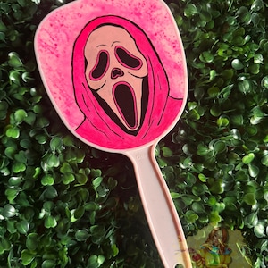 May include: A pink hand mirror featuring a Scream mask design. The mirror has a pink handle and a pink frame with the Scream mask in the center. The background is a green, leafy surface.