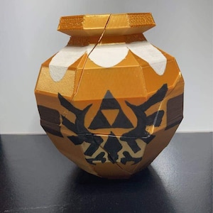 May include: A 3D printed golden pot with a black Triforce symbol and a white design. The pot is split in half and has a textured surface.