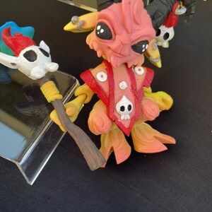 Può includere: Figurina dettagliata e colorata di una creatura con corna, una combinazione di colori rosso e arancione e un design a forma di teschio sul petto. Tiene un bastone e ha corna nere. Altre figurine sono visibili sullo sfondo.