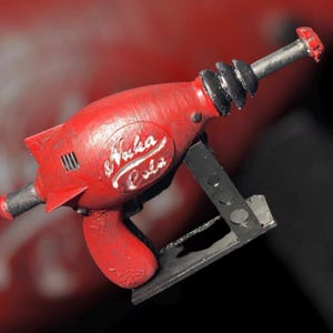 May include: A red and black toy gun shaped like a rocket with the words "Nuka Cola" printed on the side. The gun is on a black stand.