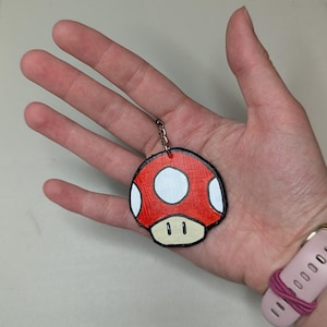 May include: A red mushroom keychain with white spots and a black outline. The keychain is held in a hand. The mushroom design is a popular video game reference. The keychain is attached to a silver chain.