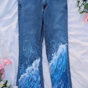 May include: Blue denim jeans with a hand-painted wave design in shades of blue and white. The jeans have a classic five-pocket design and a slight flare at the hem. White paint splatters add a unique artistic touch.