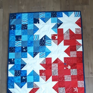Könnte beinhalten: Eine weiße, blaue und rote Patchwork-Decke mit einem Sternmuster. Die Decke besteht aus Stoffquadraten, die in einem Muster zusammengenäht sind. Die Decke ist mit einem dunkelblauen Stoff eingefasst.
