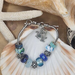 May include: A silver charm bracelet with a variety of blue and green glass beads, including a fish charm. The bracelet is on a seashell background.