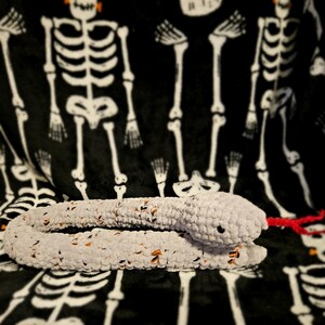 May include: A crocheted gray and orange snake with a red tongue, lying on a black and white skeleton patterned blanket.