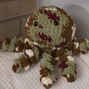 May include: A crocheted octopus plush toy with a camouflage pattern in shades of green, brown, and burgundy. The octopus has large black eyes and a white background.