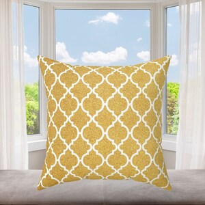 May include: Gold and white decorative throw pillow with a geometric pattern. The pillow is sitting on a light brown surface in front of a window.