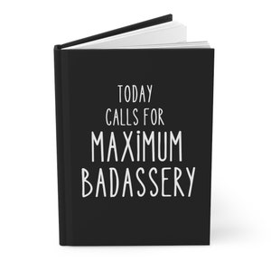 May include: A black journal with the text "TODAY CALLS FOR MAXIMUM BADASSERY" in white. The journal is open, revealing white pages. The cover is solid black, and the text is in a bold, sans-serif font.