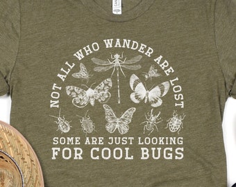 May include: Heather olive green t-shirt featuring a white insect graphic and the text "NOT ALL WHO WANDER ARE LOST SOME ARE JUST LOOKING FOR COOL BUGS". The shirt is made of a soft material.