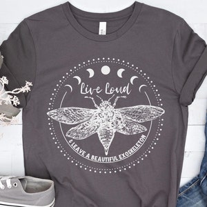 Könnte beinhalten: Ein dunkelgraues T-Shirt mit einem weißen Grafikdesign. Das Design zeigt eine Zikade mit ausgebreiteten Flügeln, umgeben von einem Kreis aus Monden und dem Text "Live Loud & Leave a Beautiful Exoskeleton".
