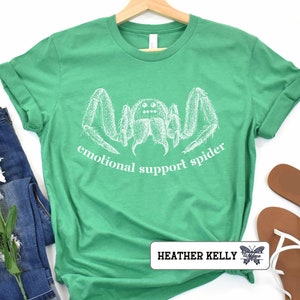 May include: A green t-shirt with a white illustration of a spider and the text "emotional support spider". The shirt is on a white background with a pair of brown sandals and a pair of blue jeans.