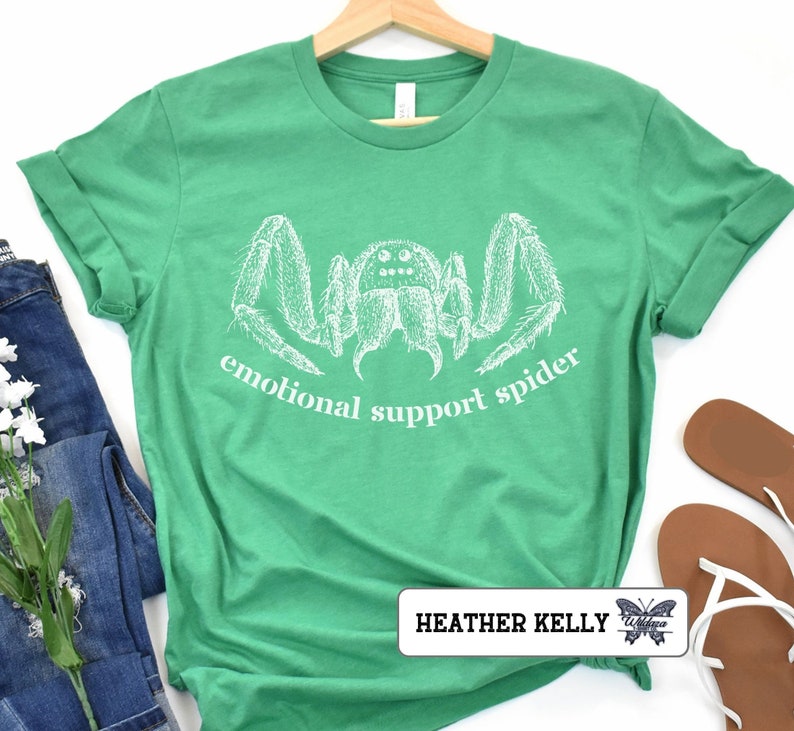 May include: A green t-shirt with a white illustration of a spider and the text "emotional support spider". The shirt is on a white background with a pair of brown sandals and a pair of blue jeans.