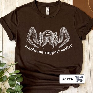 May include: A brown t-shirt with a white illustration of a spider and the text "emotional support spider".
