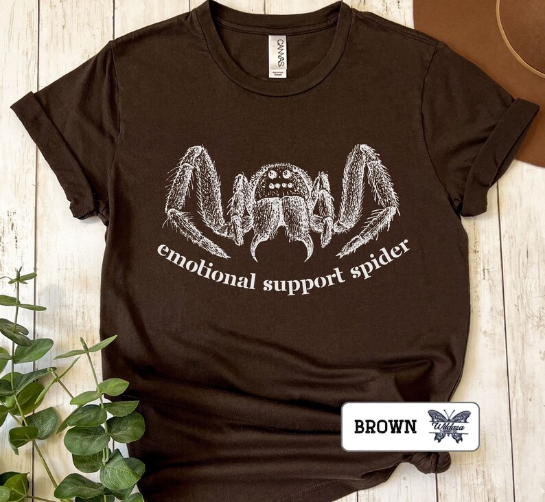 May include: A brown t-shirt with a white illustration of a spider and the text "emotional support spider".