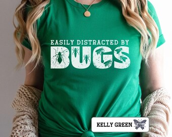 Può includere: Una maglietta verde con la scritta bianca e usurata "EASILY DISTRACTED BY BUGS". La parola "BUGS" presenta illustrazioni di insetti. La maglietta è annodata in vita. Il testo "KELLY GREEN" è in basso.