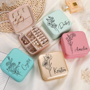 May include: Several square jewellery boxes in pastel shades, such as pink, mint green, and gold. Each box is decorated with a floral motif and a name in script. One box is open, displaying earrings, rings, and other jewellery.