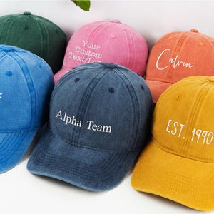 May include: A collection of colorful baseball caps in various shades, including blue, pink, orange, and yellow. Each cap features embroidered text, such as "Out Of Office," "Alpha Team," "Calvin," and "EST. 1990." The caps have a vintage, worn look.