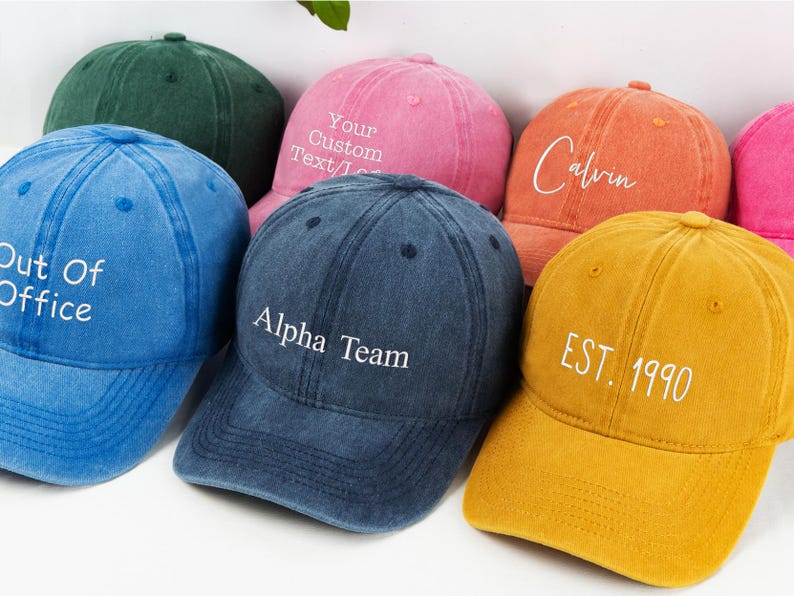 May include: A collection of colorful baseball caps in various shades, including blue, pink, orange, and yellow. Each cap features embroidered text, such as "Out Of Office," "Alpha Team," "Calvin," and "EST. 1990." The caps have a vintage, worn look.