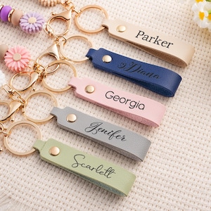 May include: A collection of personalized keychains in various colors, including beige, navy, pink, gray, and green. Each keychain features a gold-colored ring and clasp, with a name engraved on the leather strap. Decorative beaded lanyards with flower accents are also visible.