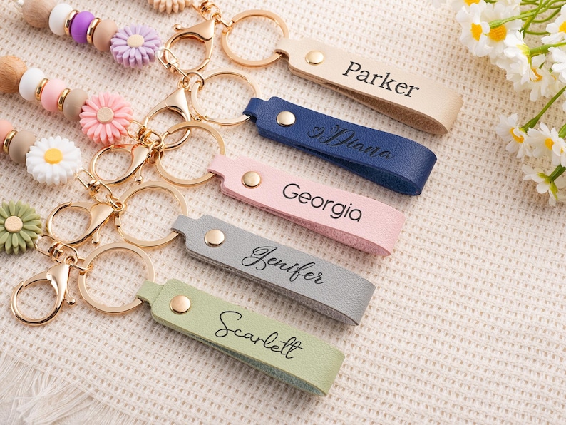 May include: A collection of personalized keychains in various colors, including beige, navy, pink, gray, and green. Each keychain features a gold-colored ring and clasp, with a name engraved on the leather strap. Decorative beaded lanyards with flower accents are also visible.