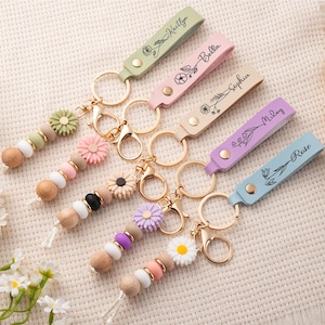 May include: A collection of personalized keychains with pastel-colored leather straps, each engraved with a name and floral design. The keychains feature wooden beads and decorative flower accents, with gold-colored hardware.