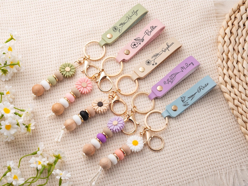 May include: A collection of personalized keychains with pastel-colored leather straps, each engraved with a name and floral design. The keychains feature wooden beads and decorative flower accents, with gold-colored hardware.