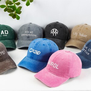May include: A collection of eight baseball caps in various colors, including green, gray, black, tan, brown, blue, and pink. Each cap features white text, with phrases like "DAD", "GRIND", and "Lily Baker".