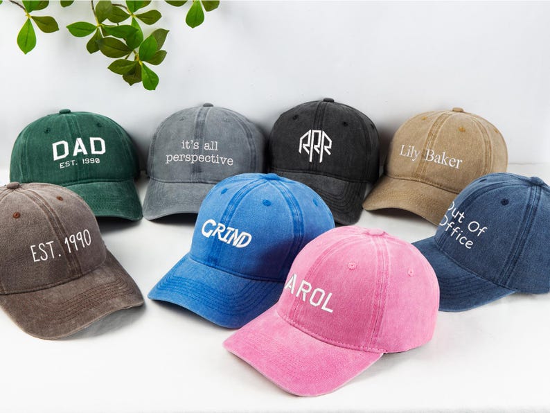 May include: A collection of eight baseball caps in various colors, including green, gray, black, tan, brown, blue, and pink. Each cap features white text, with phrases like "DAD", "GRIND", and "Lily Baker".