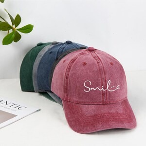 May include: A row of baseball caps in shades of green, blue, and burgundy. The burgundy cap has the word "Smile" in white script. The caps have a vintage, worn look.