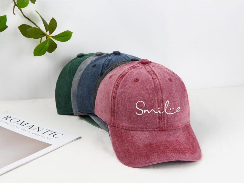 May include: A row of baseball caps in shades of green, blue, and burgundy. The burgundy cap has the word "Smile" in white script. The caps have a vintage, worn look.