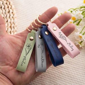 May include: A hand holds four leather keychains in green, gray, navy blue, and pink. Each keychain has a gold ring and a snap closure. The keychains are engraved with a floral design and a name: Mateo, Diana, Rose, and Miley.