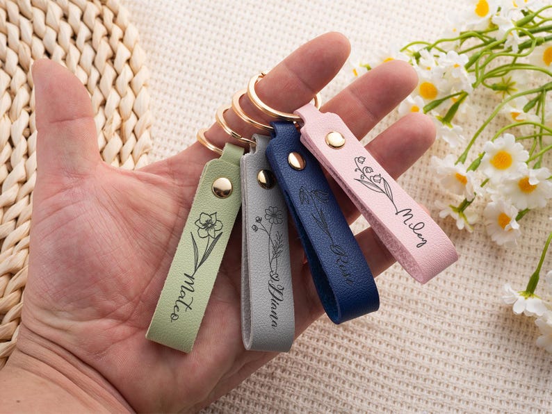 May include: A hand holds four leather keychains in green, gray, navy blue, and pink. Each keychain has a gold ring and a snap closure. The keychains are engraved with a floral design and a name: Mateo, Diana, Rose, and Miley.