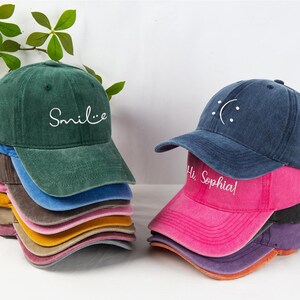 May include: A stack of colorful baseball caps in various shades, including green, blue, pink, and yellow. One stack has the word "Smile" embroidered on the front, while another has a sad face and the text "Hi Sophia".