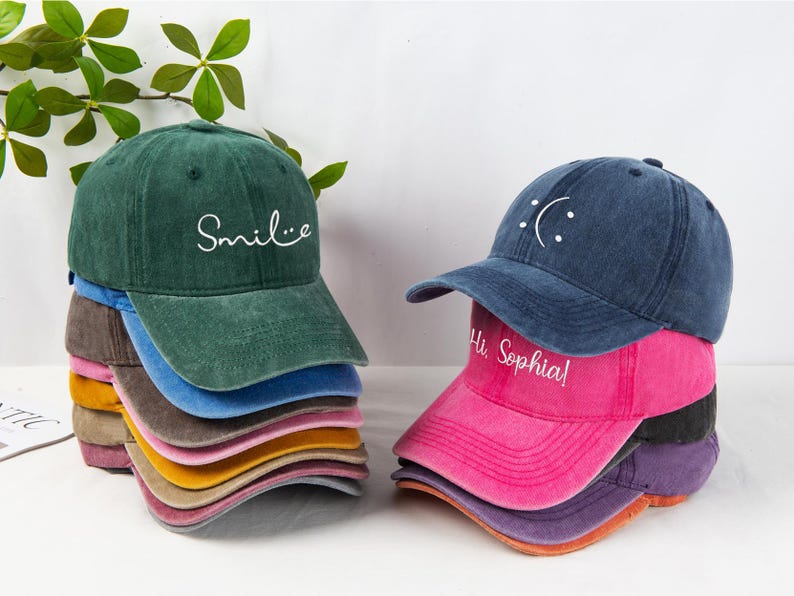 May include: A stack of colorful baseball caps in various shades, including green, blue, pink, and yellow. One stack has the word "Smile" embroidered on the front, while another has a sad face and the text "Hi Sophia".