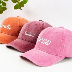 May include: Three baseball caps in shades of orange, burgundy, and pink. Each cap has a curved brim and a top button. The orange cap reads "Smith", the burgundy cap reads "ly Baker", and the pink cap reads "GRIND" in white lettering.